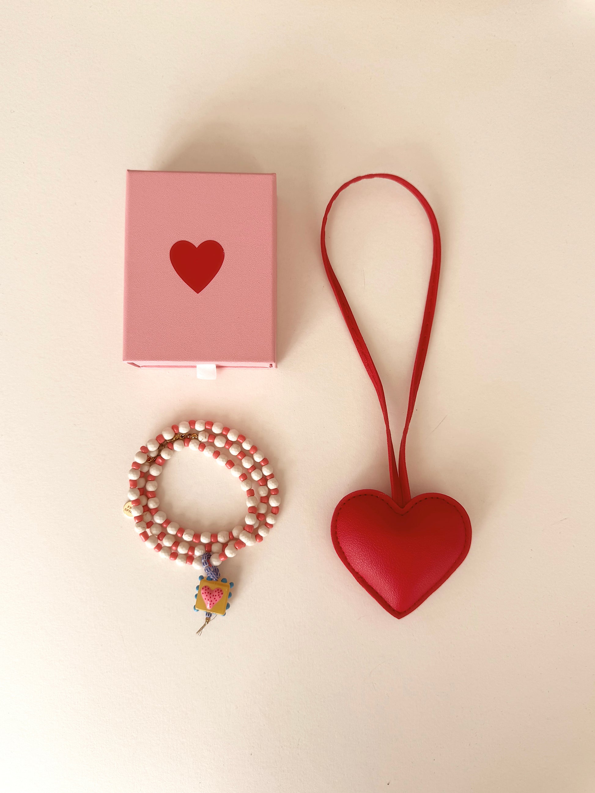 Red heart-shaped keychain, pink box with heart, and beaded bracelet on a beige background